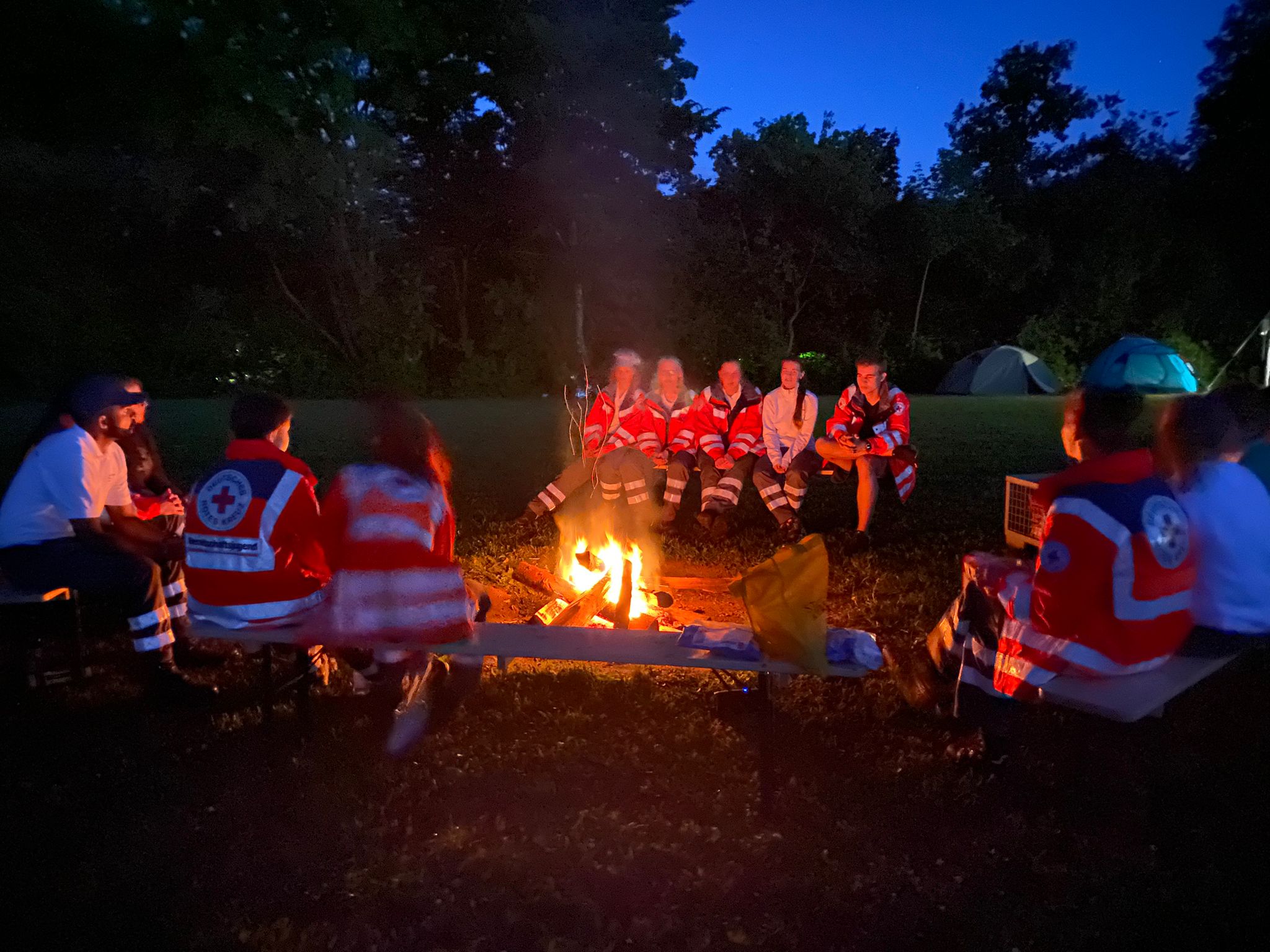 Jugendliche und Betreuer in Rotkreuz-Jacken sitzen um ein Lagerfeuer. Im Hintergrund sind vor Nachthimmel Laubbäume zu erkennen.